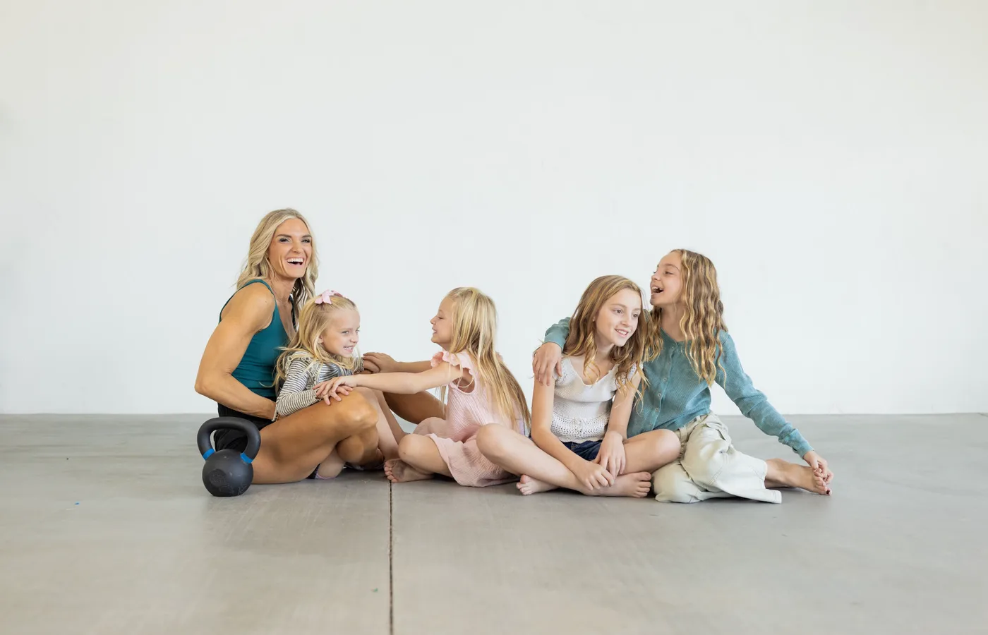 Andrea Allen sitting on a studio floor with her four girls, all laughing together, a kettlebell beside her