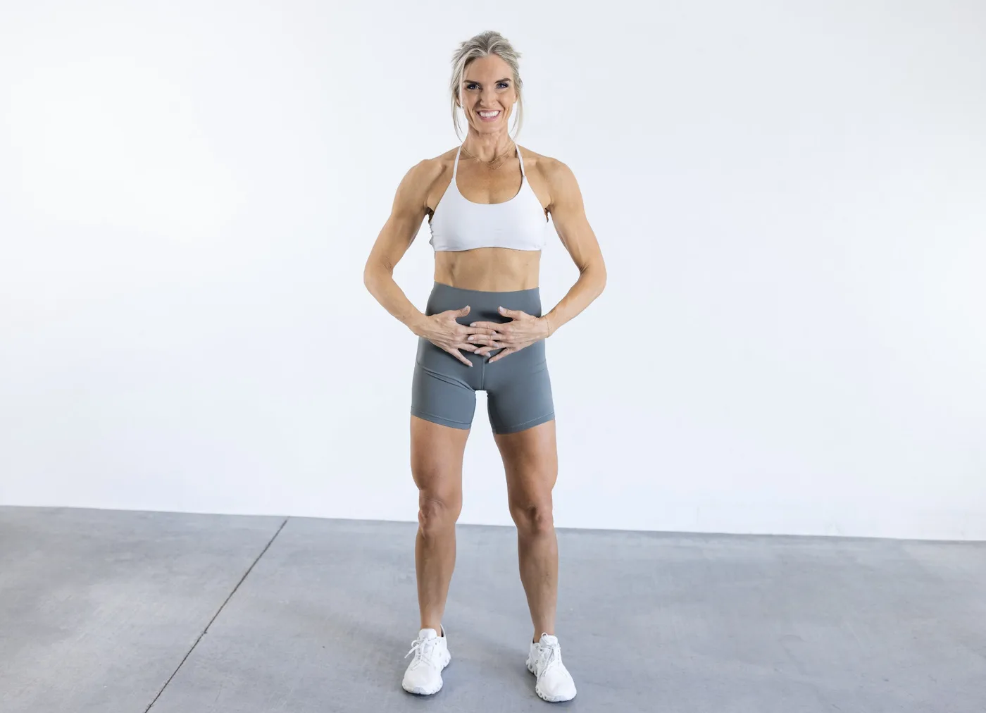 Andrea standing with both hands on her lower abdomen, demonstrating diaphragmatic breathing and deep-core connection